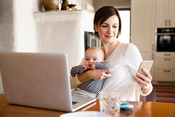 mother holding a child and a phone in front of a laptop computer